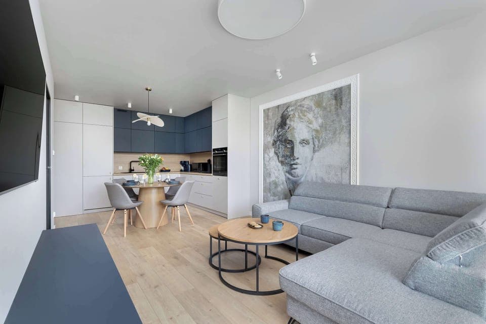 A bright lounge area with a gray corner sofa, a circular table, and natural views through the windows.