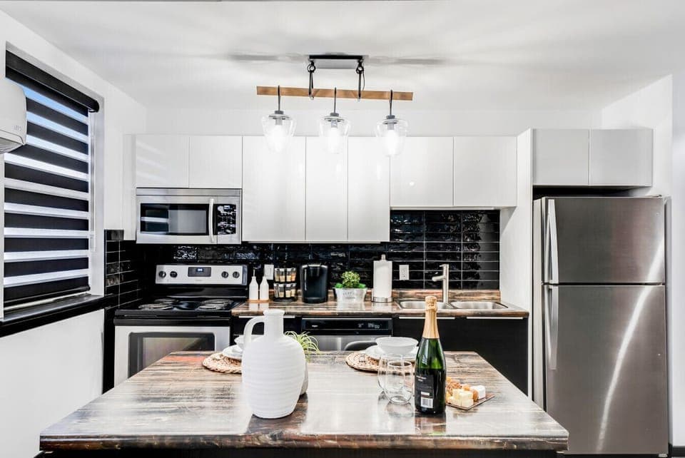 Fully equipped modern kitchen with sleek white cabinets, black subway tile backsplash, and stainless steel appliances including a fridge and microwave. Features a spacious island with seating, perfect for dining and entertaining. Ideal for culinary needs!