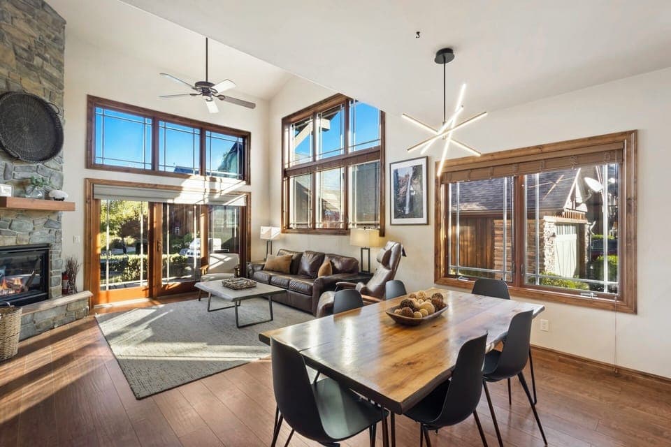 Open living room and dining room bosting cathedral ceilings, an abundance of windows, a fireplace, a smart TV and a large dining room table.