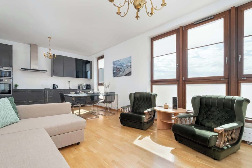 A living room with a cozy sofa, armchairs, and TV area. The wooden floor and light walls create a warm, inviting atmosphere.