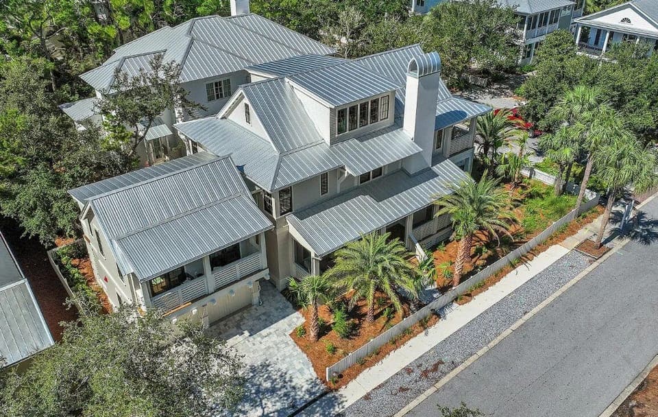 Aerial view of a beautiful coastal home with metal roofing and tropical landscaping in a tree-lined neighborhood.