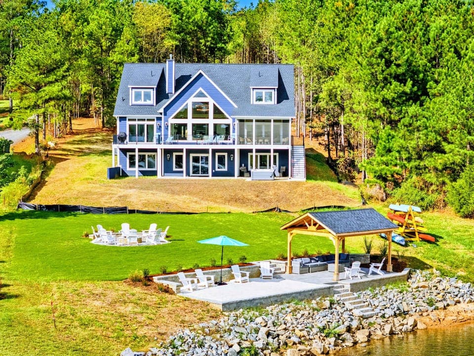 Home Exterior - The striking blue lakefront home from the water's edge, with the patio, pavilion, and fire pit circle in the foreground.