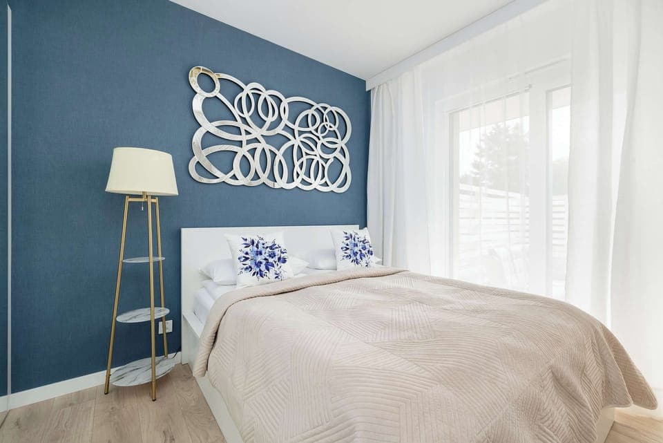 A modern bedroom with a blue accent wall and stylish decor above the bed. A soft beige blanket and elegant lamp add warmth to the space.