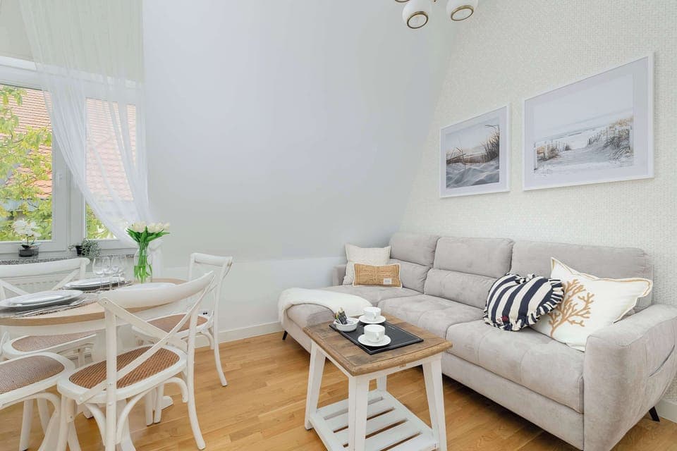 A bright living room with a light sofa and a round glass table. White walls and natural light create an airy atmosphere.