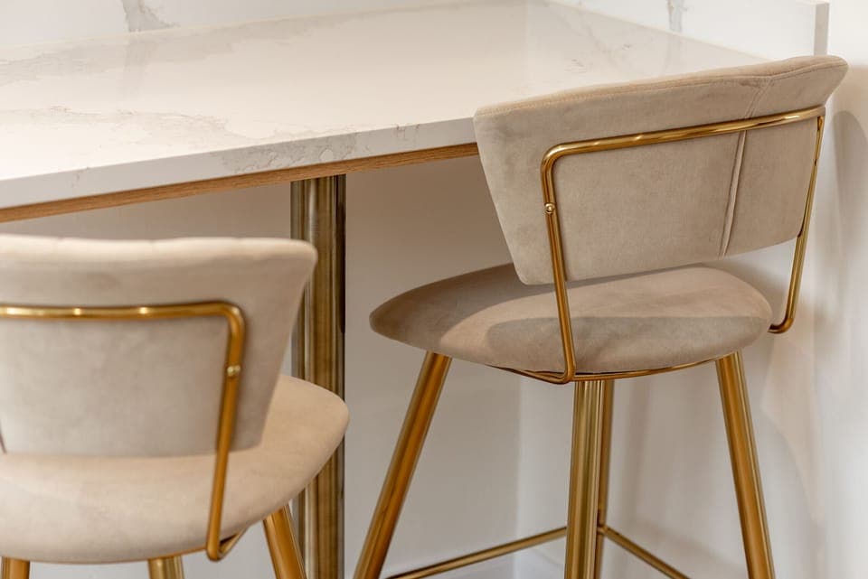 Close-up of a kitchen breakfast bar with plush beige velvet stools featuring gold frames and legs.