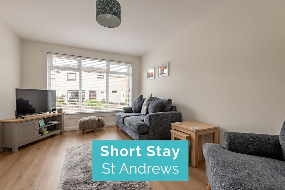 Spacious living room with large front-facing window, grey sofa, TV unit, and decorative highland cow footstool.