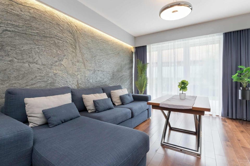 A stylish living room with dark gray sofas, wooden flooring, and large windows covered by sheer curtains. Warm ceiling lighting adds a modern touch.