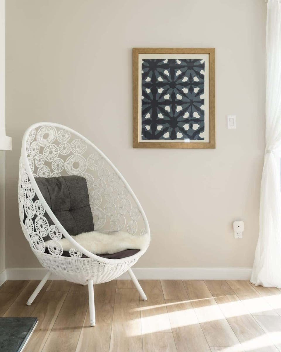 The intricate white wicker design, paired with the dark cushion and faux fur accent, makes it look both cozy and stylish. It’s the perfect reading nook or relaxation spot. The framed artwork adds a nice contrast. Such a well-curated corner!