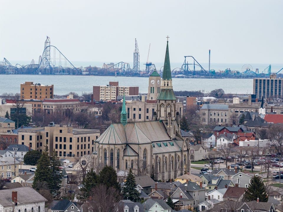 Historic cityscape featuring a prominent church with green spires, surrounded by diverse architecture and waterfront amusement park rides in the distance.