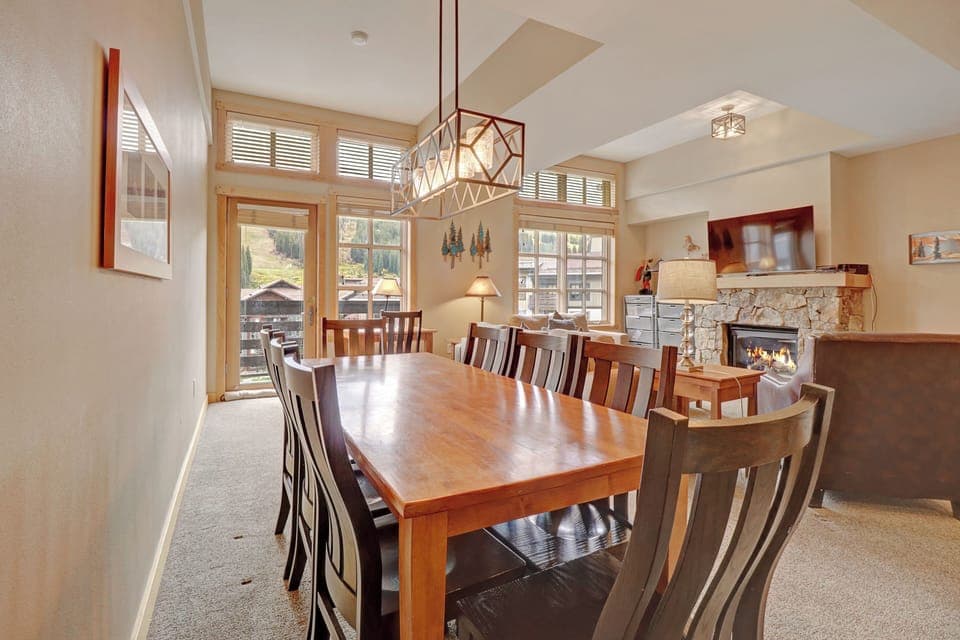 Spacious dining area featuring a large wooden table for eight, illuminated by a contemporary chandelier. Enjoy mountain views through expansive windows and the cozy ambiance of the nearby stone fireplace.