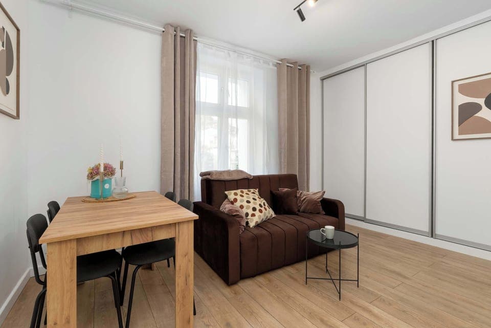 A cozy living room area with a brown sofa and a small wooden table, next to a window with light curtains. The room also features a workspace with a wooden desk and chair.