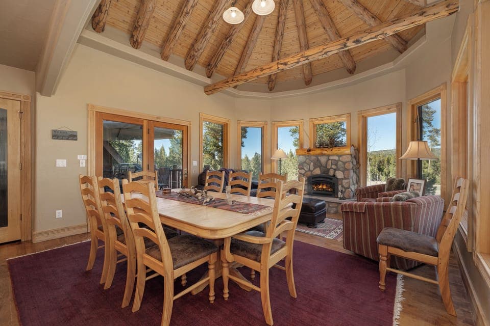 Gather around this rustic dining table beneath soaring wood beams, while the stone fireplace creates warmth and panoramic windows frame nature's beauty.
