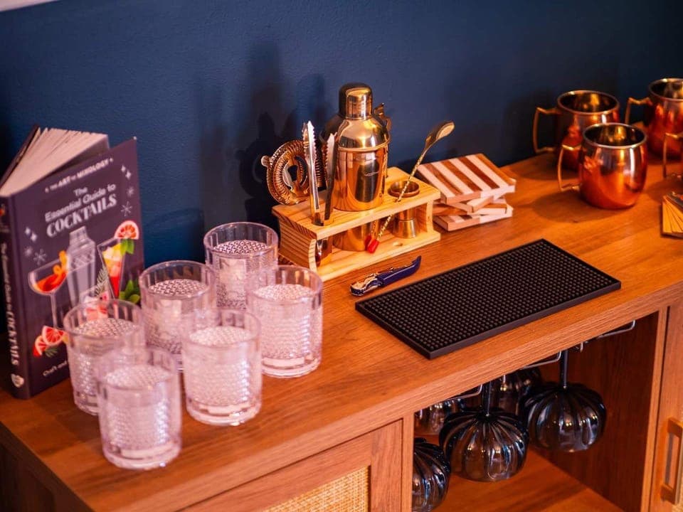 Shake up some fun! This custom bar features a sleek teal backdrop, a gold shaker set, and crystal glassware. Complete with copper mugs and a cocktail guide—it's the perfect spot to toast your stay! 🥂🍸✨