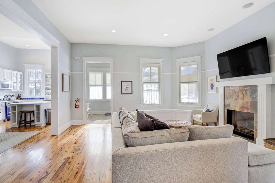 A bright living room with a stone fireplace, mounted Smart TV, and wraparound windows creates a relaxing space to gather after a day outdoors.