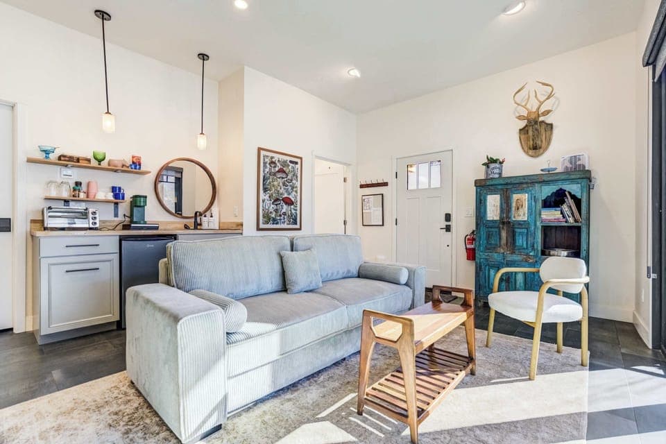 A cozy arrangement of the sofa, accent chair, wooden coffee table, blue cabinet, and visible kitchenette creates a warm gathering space framed by sliding glass doors facing the mountains.