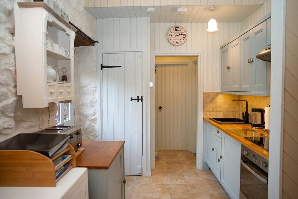 Narrow kitchen with blue cabinets, wooden worktop and sink, with a doorway and wall clock