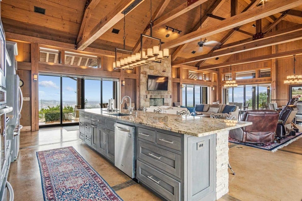 An open-concept living space that seamlessly blends a chef's kitchen with a cozy lounge area, highlighted by soaring wood-beamed ceilings and a stunning stone fireplace create perfect setting while soaking in breathtaking panoramic views.