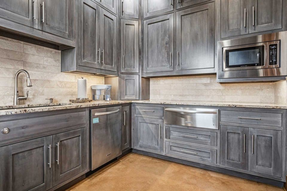 This stunning kitchen features a distinctive grey wash finish on its custom-built cabinets, creating a rustic-chic aesthetic. The shaker-style doors add a classic touch, while the modern stainless steel appliances bring a sleek, contemporary element.