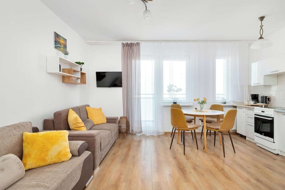 A bright living area with a sofa, a dining table with chairs, and a kitchenette in the background. The space is cozy and well-lit, ideal for relaxing or dining.