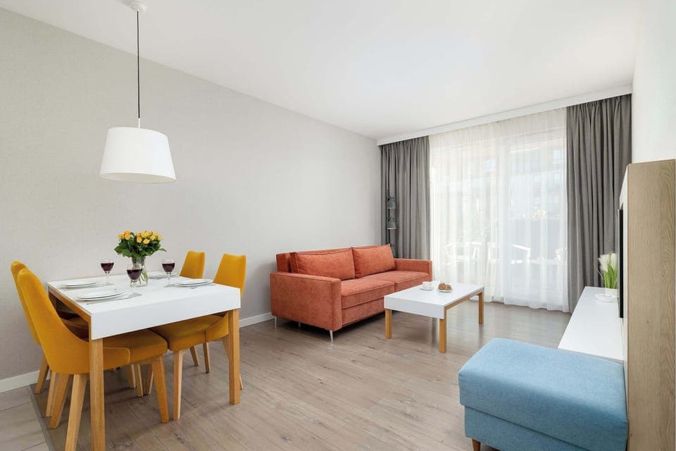 A bright living and dining area with a wooden table, mustard-yellow chairs, a salmon-colored sofa, and large curtains letting in daylight.