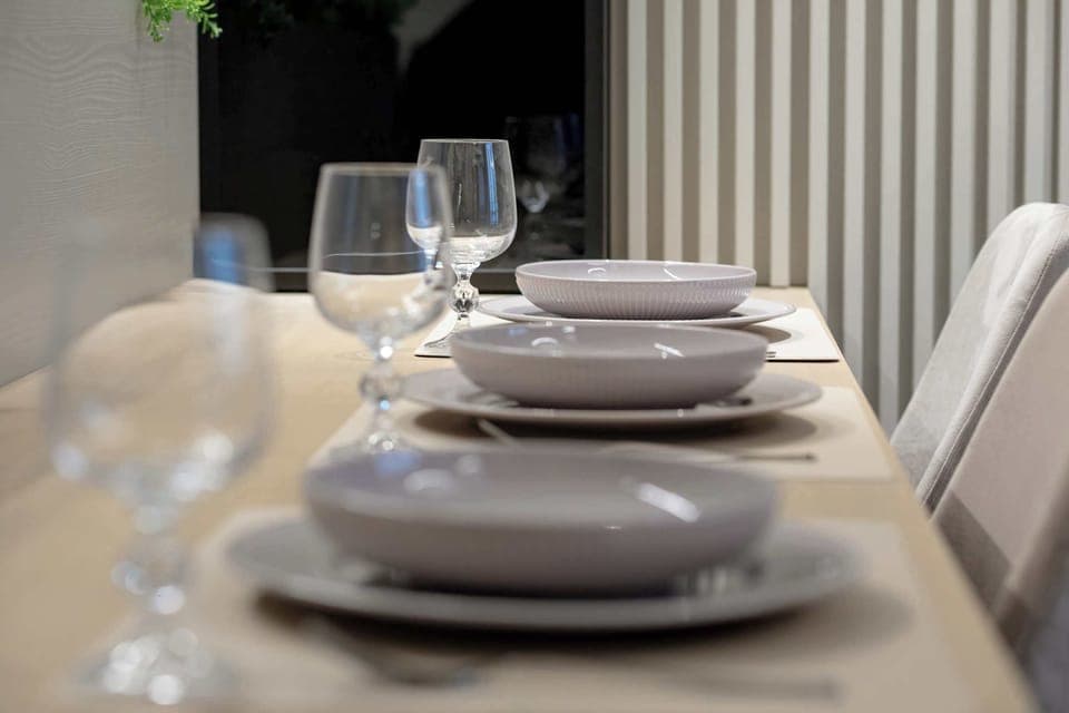 A close-up of stacked plates and glassware arranged neatly on a dining table. The setup suggests a prepared dining experience. Soft lighting highlights the clean presentation.