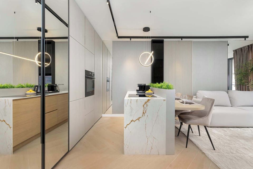 An open-plan kitchen and dining area with a central island. Light cabinetry, marble-style surfaces, and pendant lighting define the modern design. The layout feels spacious and functional.
