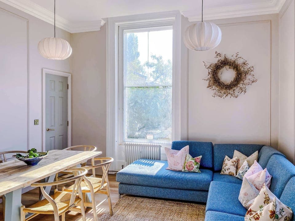 Sunlit living and dining space with a cozy blue sofa, decorative pillows, and a woven rug. A wooden table with fruit centerpiece sits beneath pendant lights, while a wall wreath adds charm. Warm, inviting, perfect for relaxing or entertaining.