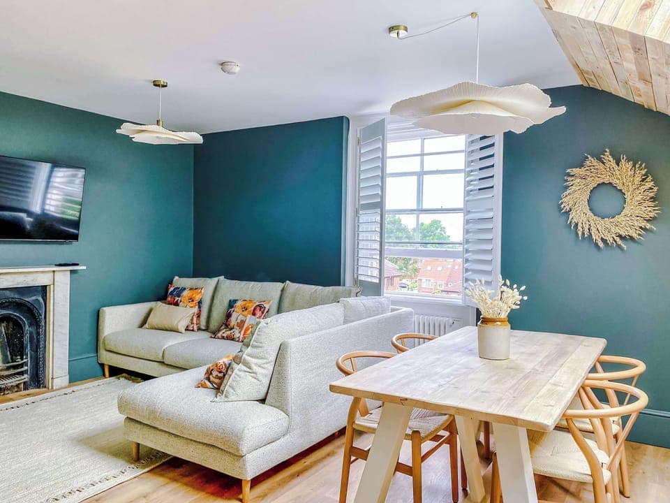 Living room with a light gray sectional sofa and decorative pillows, set beside a sleek fireplace and wall-mounted TV. Muted green walls, wood flooring, and pendant lights create a cozy yet modern space that flows seamlessly into the dining area.
