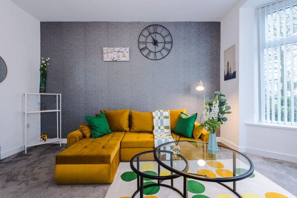 Living room with a mustard corner sofa, round glass coffee table, wall clock, and large window with vertical blinds.