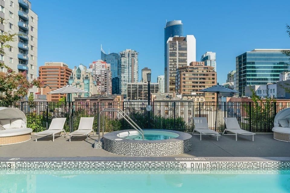 Set against a striking city skyline, the rooftop pool area features a relaxing hot tub and lounge chairs, inviting guests to unwind in a vibrant atmosphere.