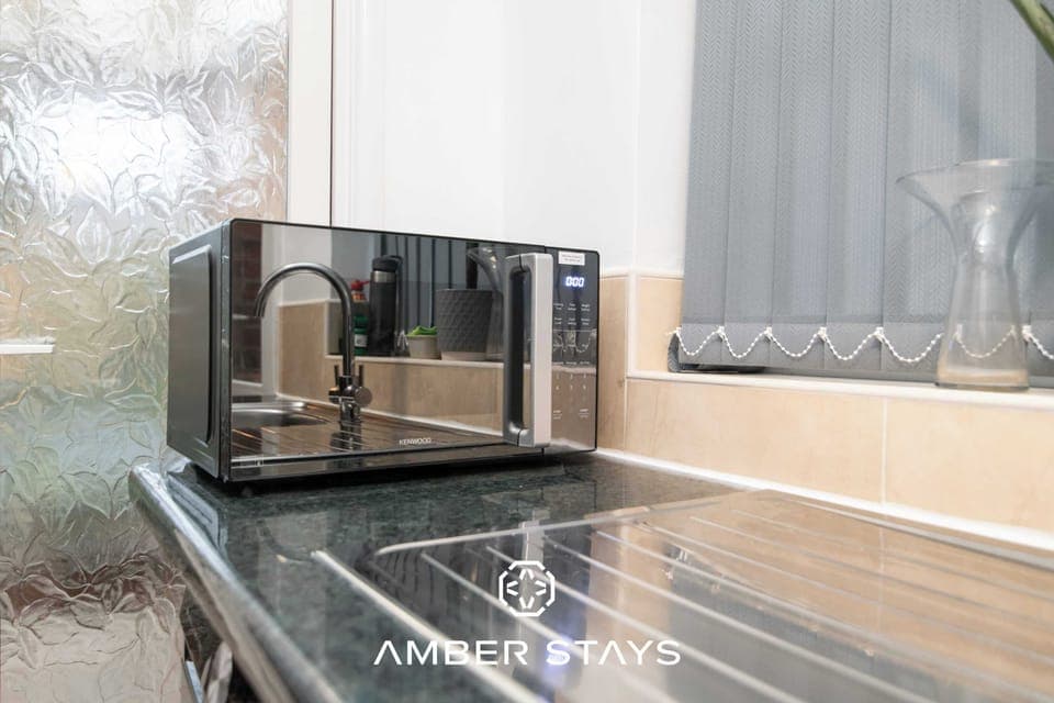 A sleek, mirrored microwave sits on a dark countertop, reflecting the surrounding kitchen and a window with grey blinds.