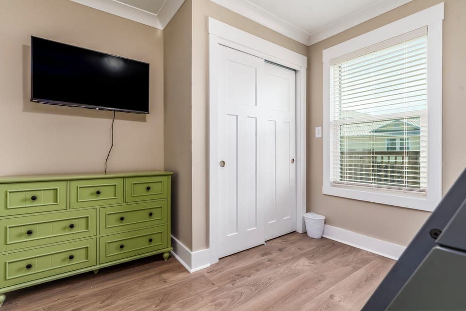 Your peaceful retreat features a wall-mounted TV and charming green dresser, with natural light streaming through blinds to create a cozy atmosphere.