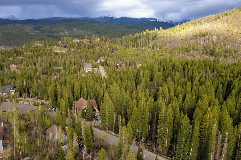 Aerial view of the property nestled among dense evergreen forests, with snow-capped mountain peaks rising in the distance and scattered vacation homes throughout the wooded valley.