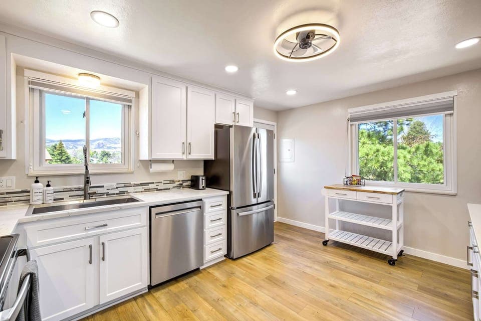 A second angle reveals the full kitchen layout, with stainless refrigerator, stove, and ample counter space along both walls. Large windows on either side bring in natural light and frame views of the backyard and surrounding neighborhood.