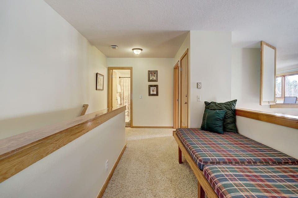 A hallway with carpeted flooring, wooden trim, and a ceiling light leads to various rooms. A cushioned bench with green and plaid pillows is placed along a wall on the right side.