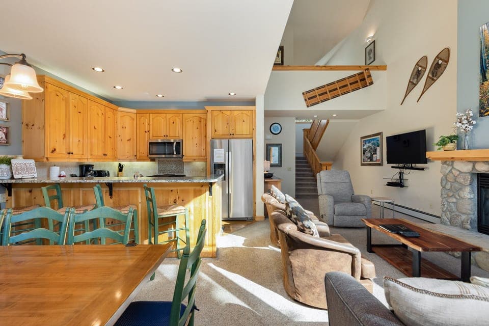 A cozy, well-lit living room and kitchen with wooden cabinets, a stone fireplace, a mounted TV, and seating area. Stairs lead to an upper floor. Decor includes wall-mounted skis.