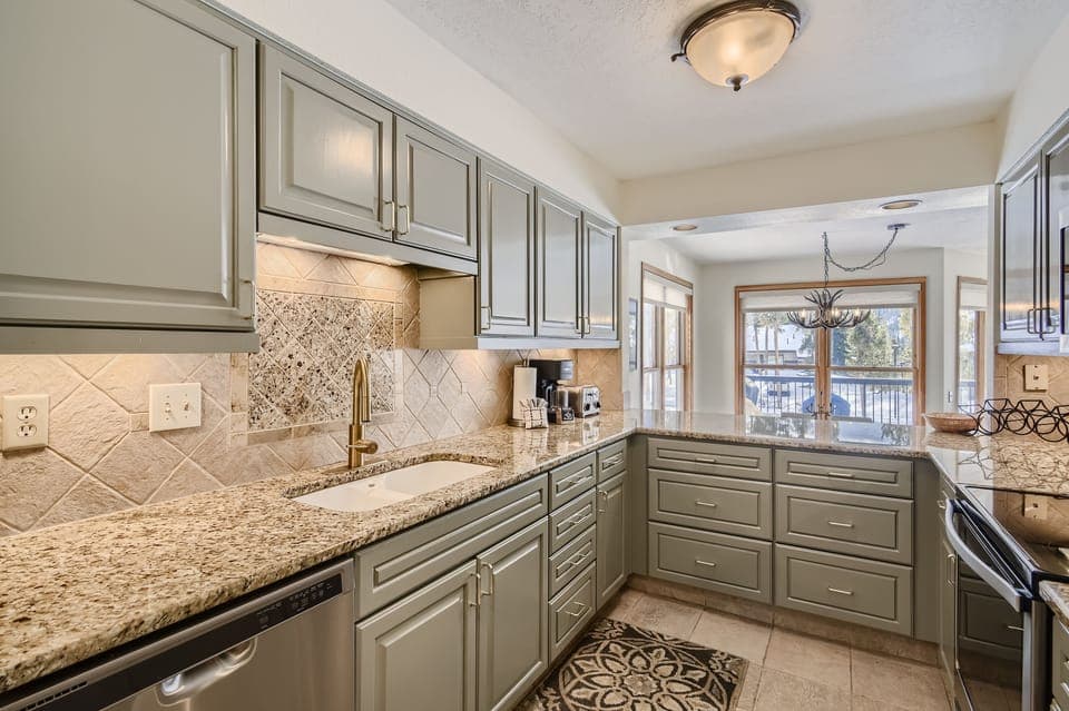 A modern kitchen with granite countertops, green cabinetry, stainless steel appliances, and a tile backsplash. A windowed dining area with a chandelier is visible in the background.