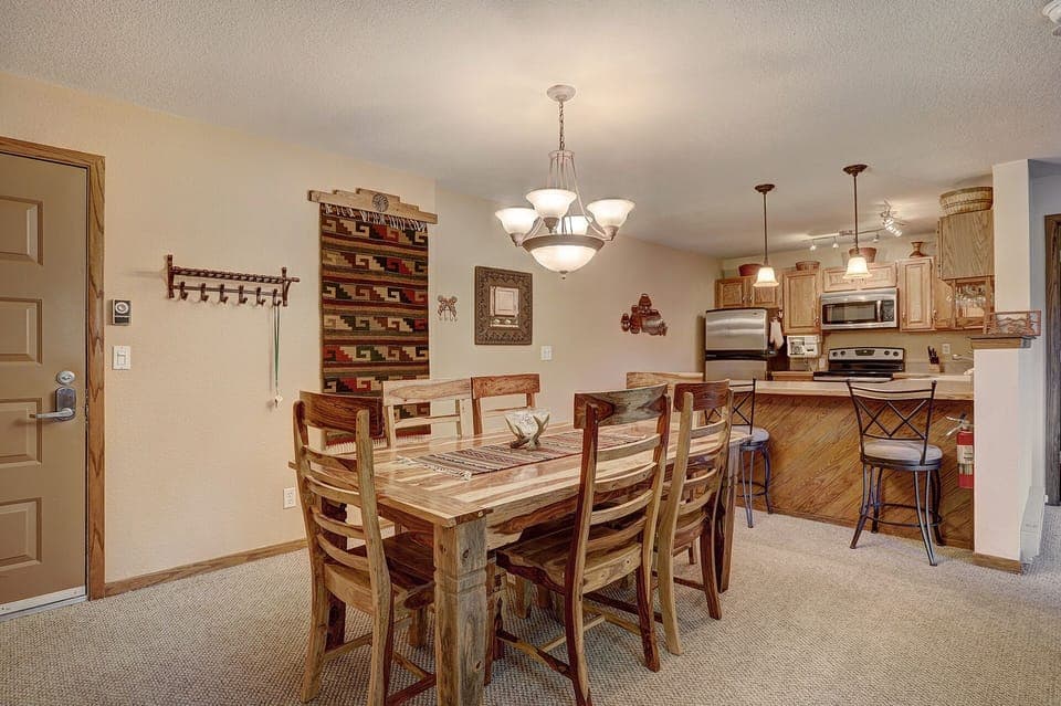 A cozy dining area with a rustic wooden table and six chairs, adjacent to a kitchen with wooden cabinets, stainless steel appliances, and a breakfast bar with three stools. Warm lighting illuminates the space.