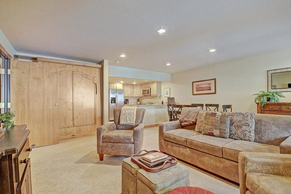 A cozy living room with beige furniture, a wooden armoire, and a flat-screen TV. The space includes a small kitchen area with stainless steel appliances and a dining table in the background.
