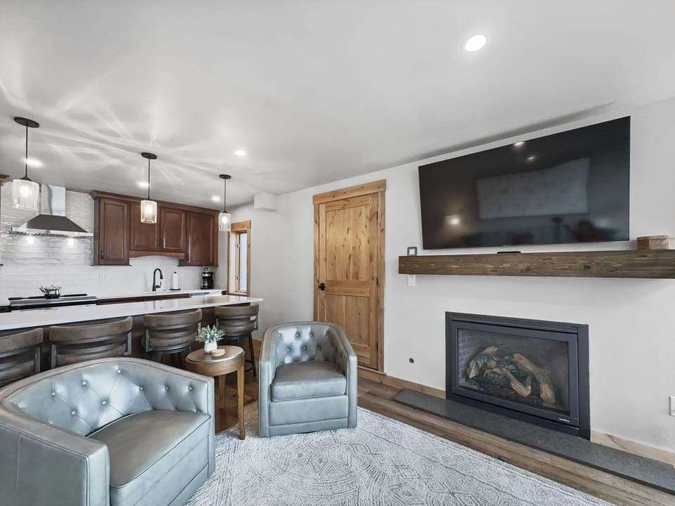 A modern living room with two gray armchairs, a wooden coffee table, a wall-mounted TV above a fireplace, and a view into the kitchen with dark cabinets and pendant lights over a countertop.