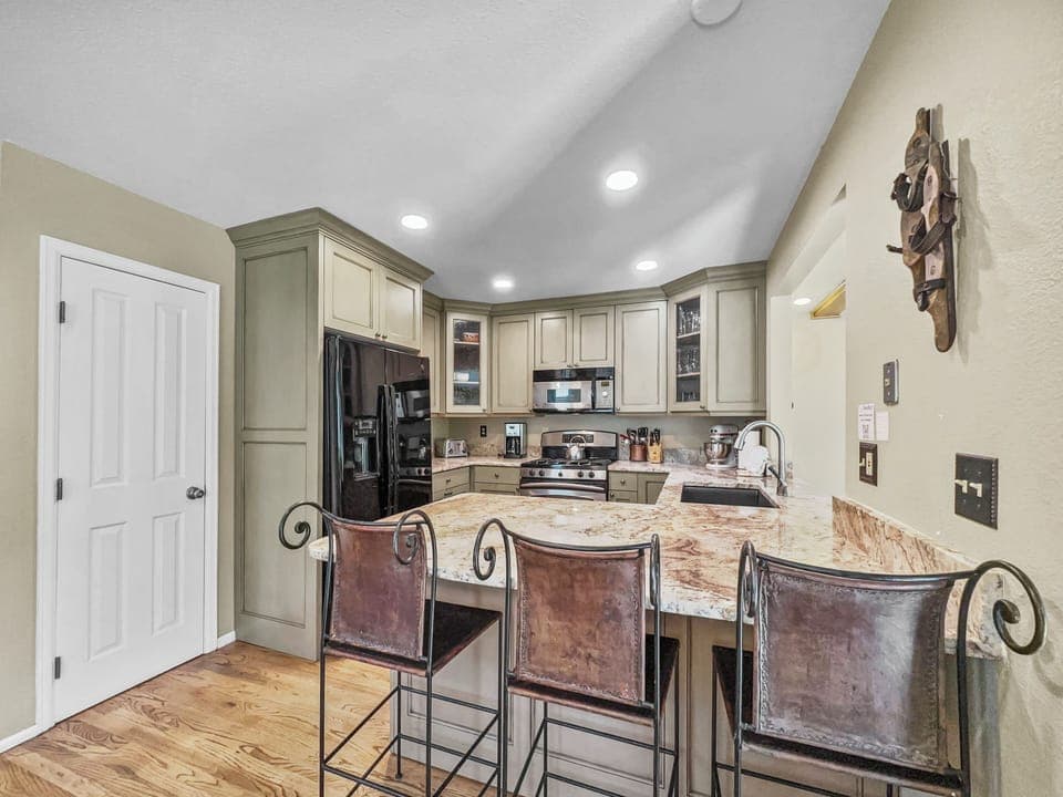 A modern kitchen with green cabinets, a granite countertop, stainless steel appliances, a black refrigerator, and three high-back leather bar stools. There is a door on the left and wood flooring.