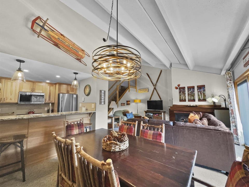 A cozy living and dining area with wooden furniture. The modern kitchen is in the background. Decor includes a chandelier, a sled hanging on the wall, and a fireplace with skis mounted above it.