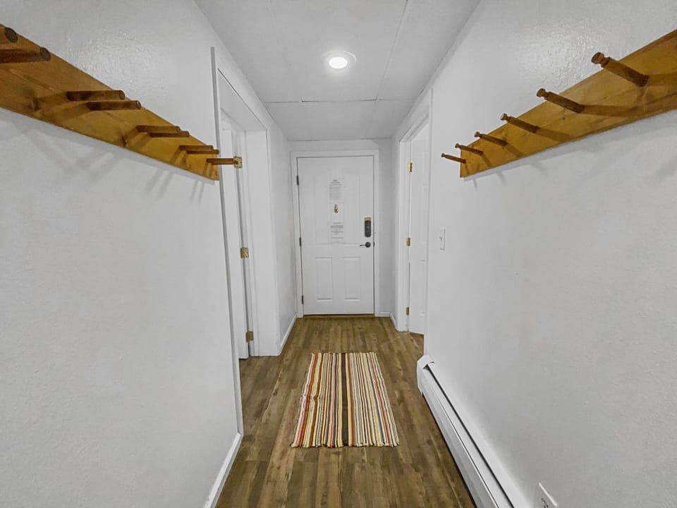 A narrow hallway with white walls, wooden coat pegs on both sides, a white door at the end, and a colorful striped rug on a wooden floor.