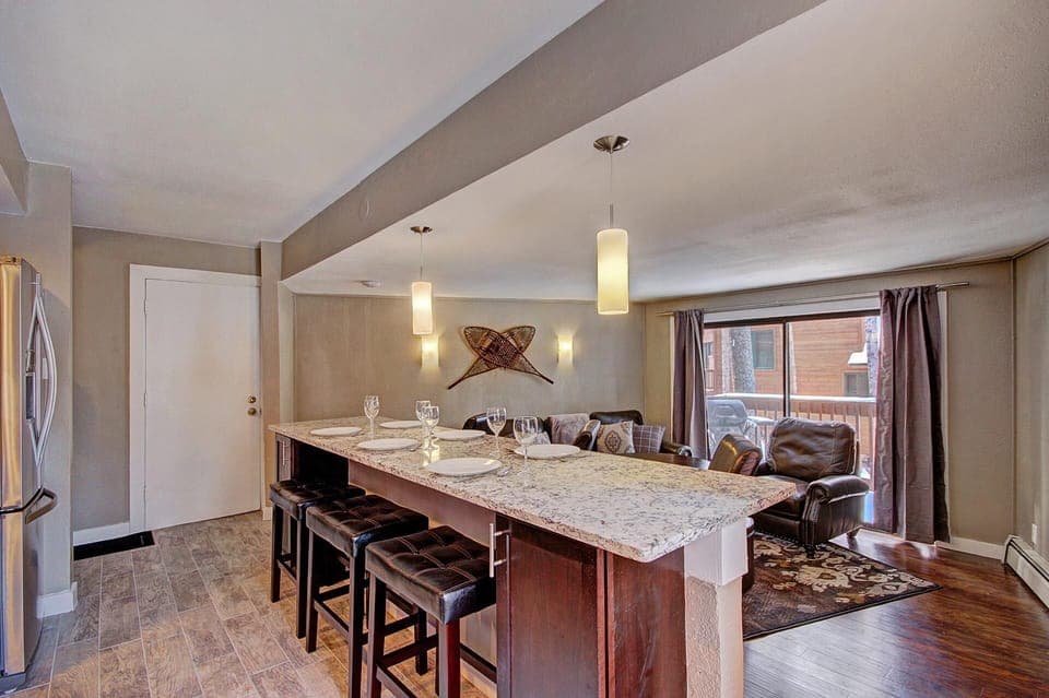 A modern kitchen with a marble island, bar stools, pendant lights, and place settings, adjoining a living area with leather sofas and large windows.
