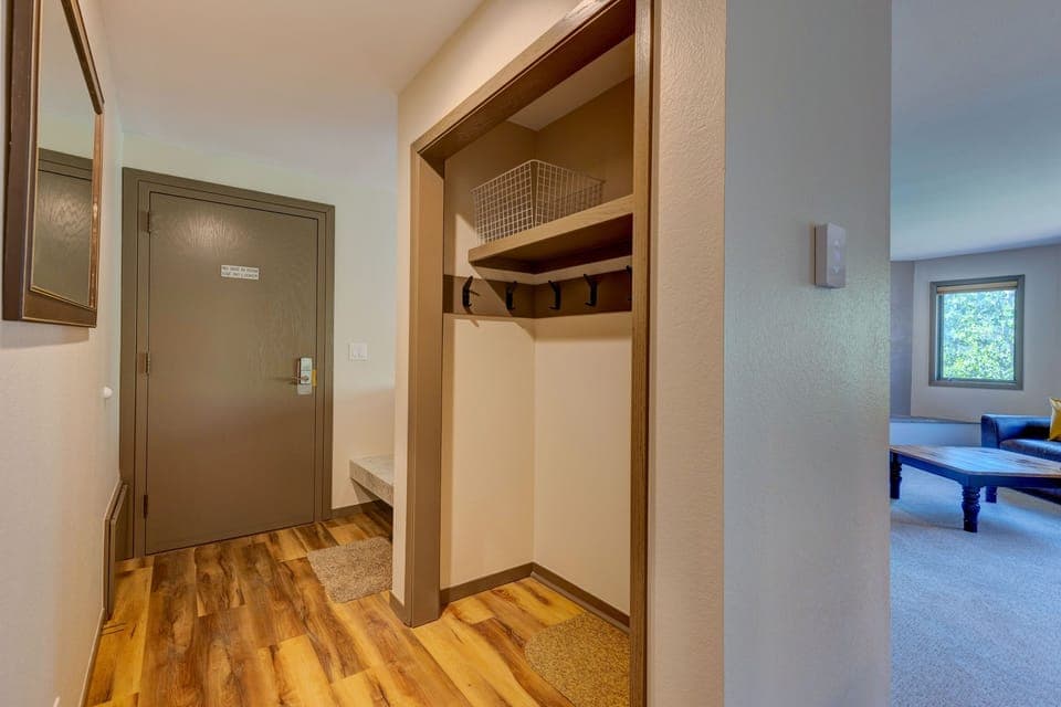 Entryway with wooden floor, wall hooks for hanging items, a shelf with a basket, and open doorway to a spacious, carpeted living room. Door at left has a door handle and small sign.
