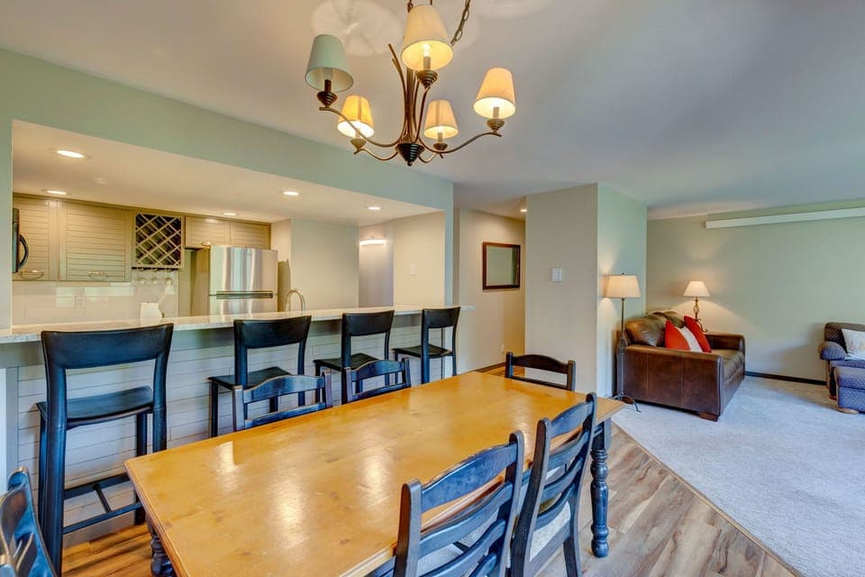 A modern open-concept dining and living area with a wooden dining table, black chairs, overhead chandelier, bar seating, kitchen with stainless steel appliances, and a seating area with a couch and lamp.