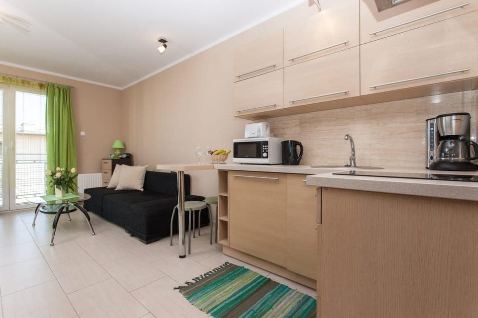 A view of the kitchen and dining area, with a small dining table for two and bright green curtains hanging by the large window, which brings in plenty of natural light.