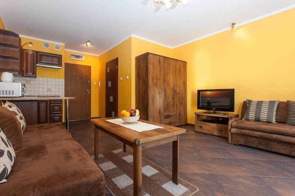 A living room with warm yellow walls, featuring a brown sofa, armchairs, and a coffee table. In the background, there's a dining area and a kitchen, creating an open-plan layout.