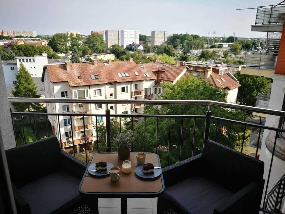 A small balcony with a table and two chairs, offering a scenic view of the city skyline, including rooftops and trees in the distance.