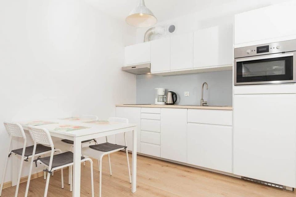 Dining area with white table and white chairs next to kitchenette with white furniture.
